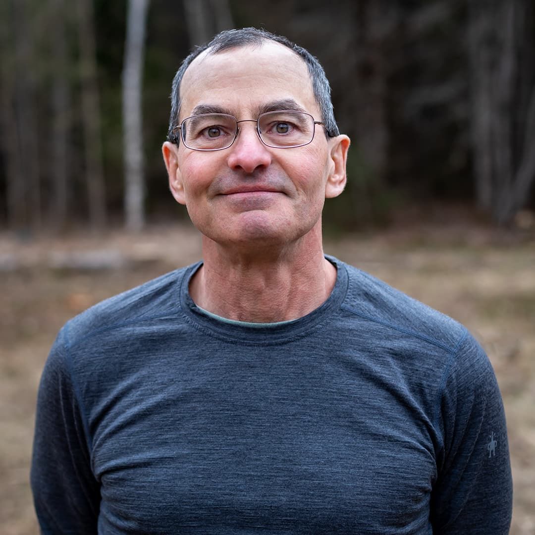 Smiling person in a gray shirt standing outdoors with trees blurred in the background