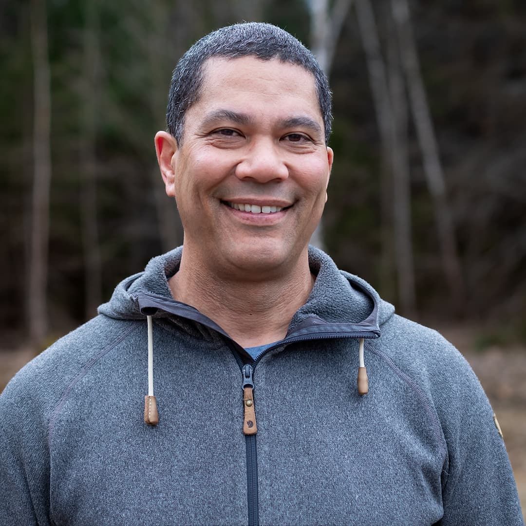 Smiling person in a gray hoodie outdoors with blurred trees in the background