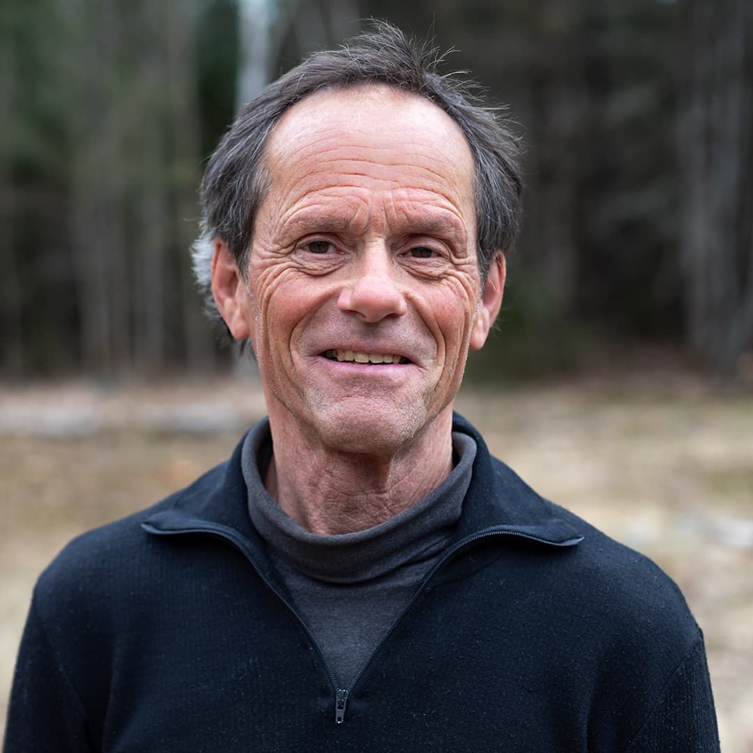 Smiling man in a dark jacket standing outdoors in a wooded area