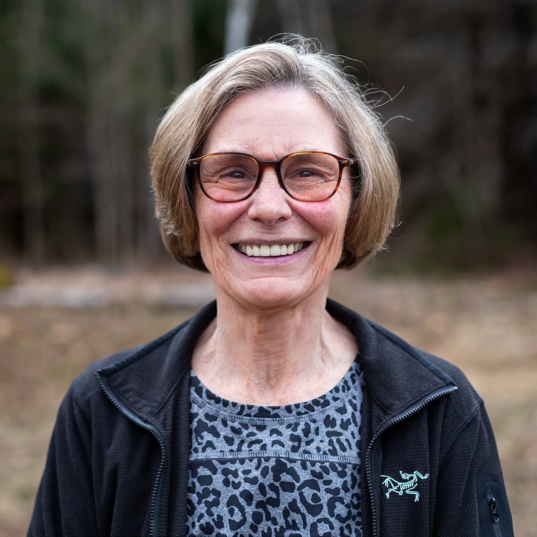 Smiling woman with glasses and short gray hair, wearing a black jacket outdoors
