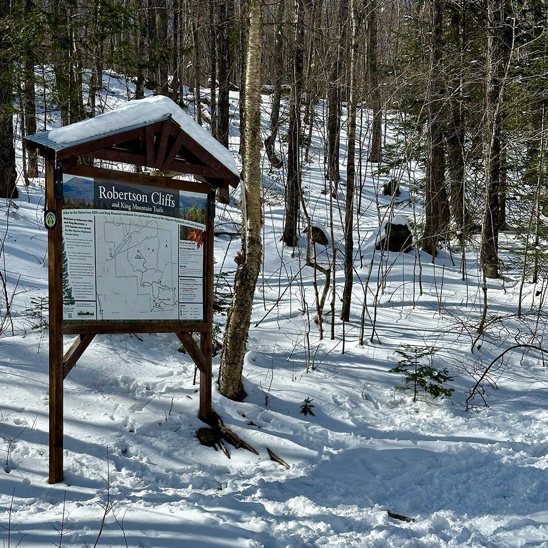 Snowy forest trailhead sign beside a wooded path, with snow covering the ground and trees.