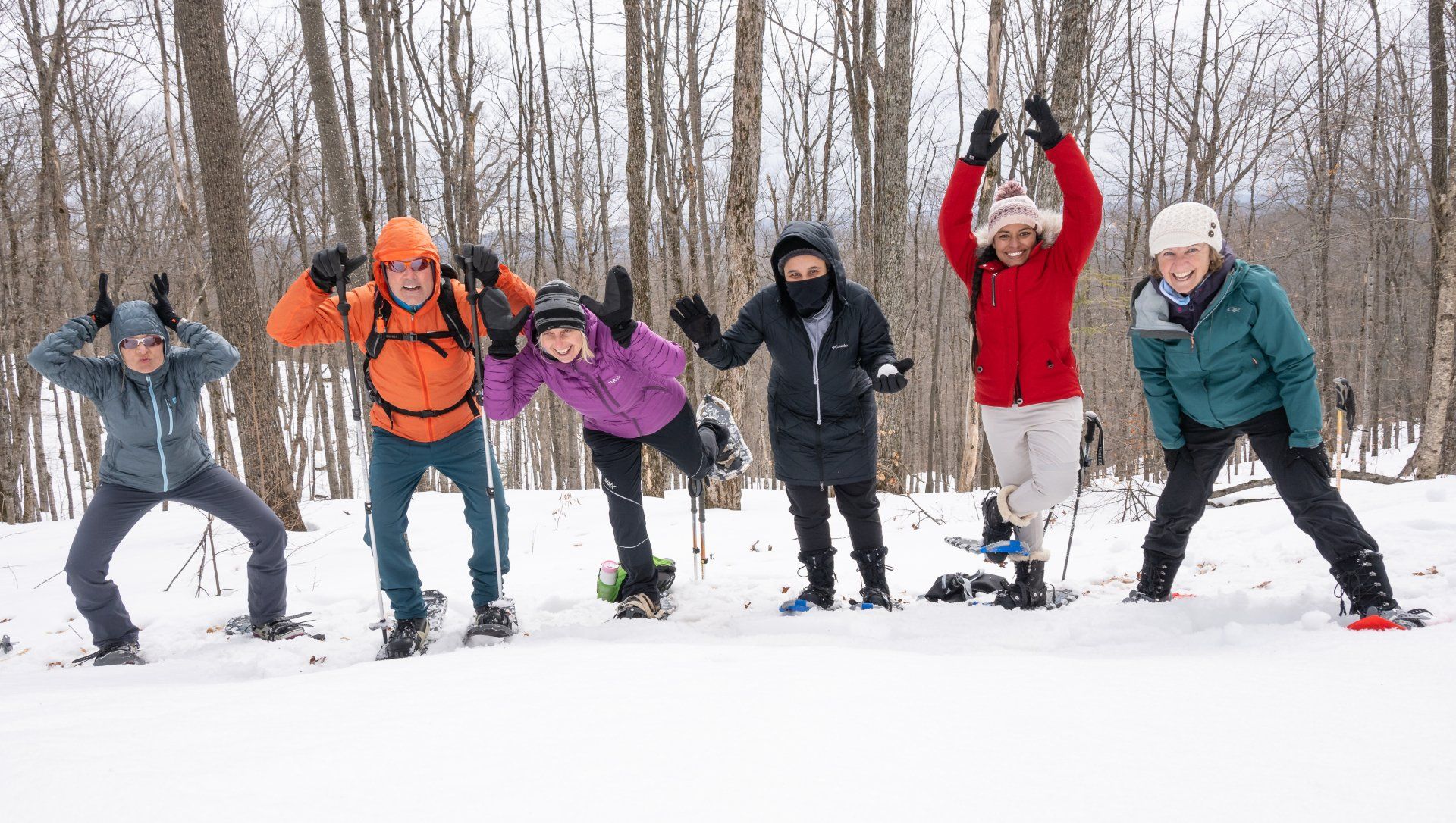 Six people in winter gear pose playfully in snow-covered woods; some with arms raised, snowshoes visible.