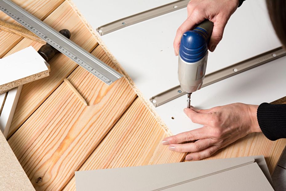 Mani che usano un trapano su una lavagna bianca accanto a righelli di legno e metallo impilati.