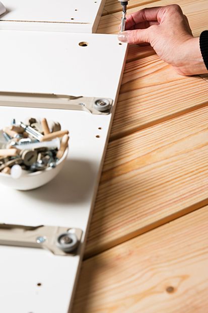 Mano che inserisce una vite in un pannello bianco di un mobile su una superficie di legno, con gli attrezzi nelle vicinanze.
