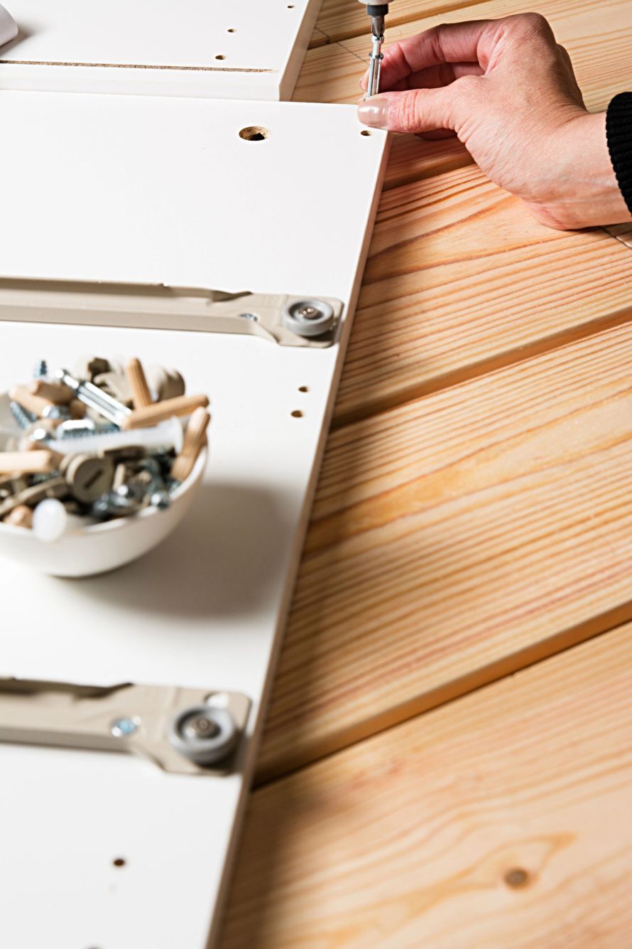 Mano che inserisce una vite in un pannello bianco di un mobile su una superficie di legno, con gli attrezzi nelle vicinanze.