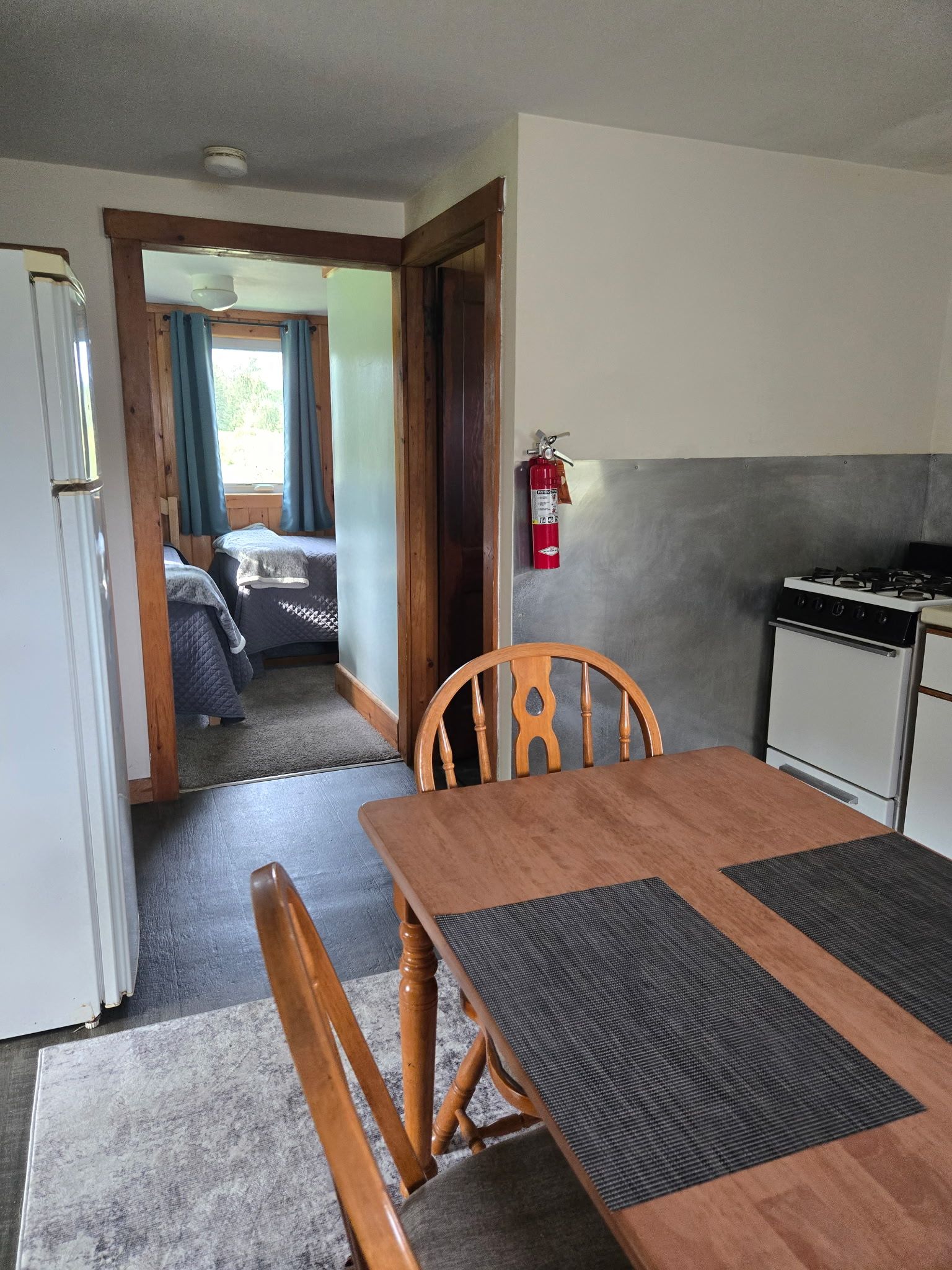 Small kitchen with wooden table, white refrigerator, stove, and doorway to a bedroom in the background