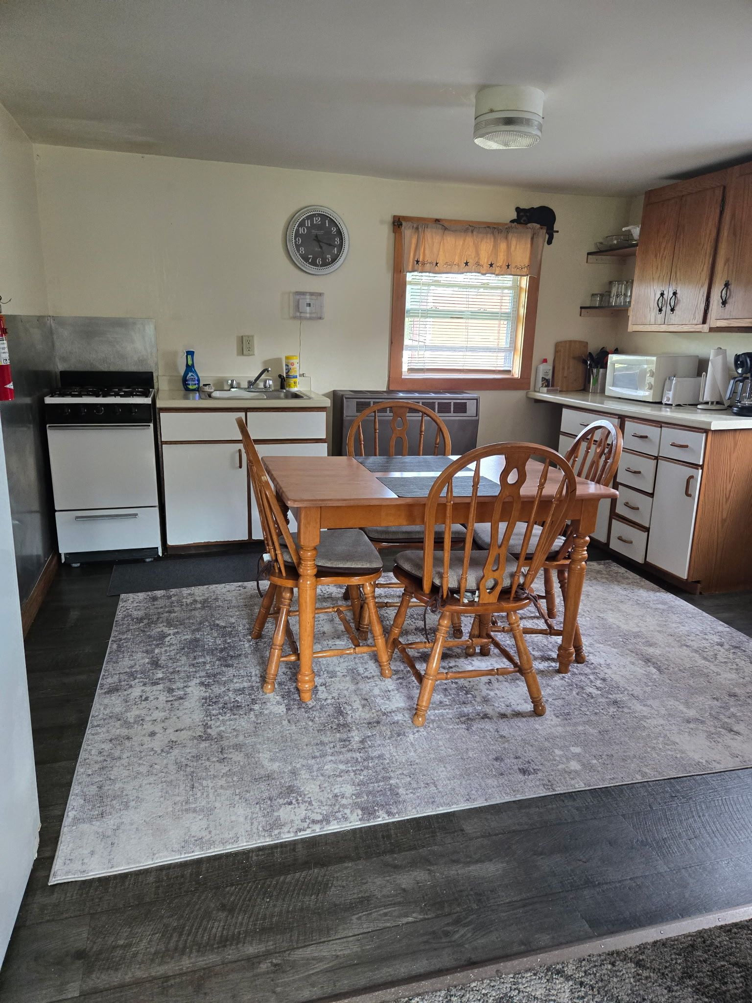 Small kitchen with wooden table, chairs, cabinets, appliances, and a patterned rug.