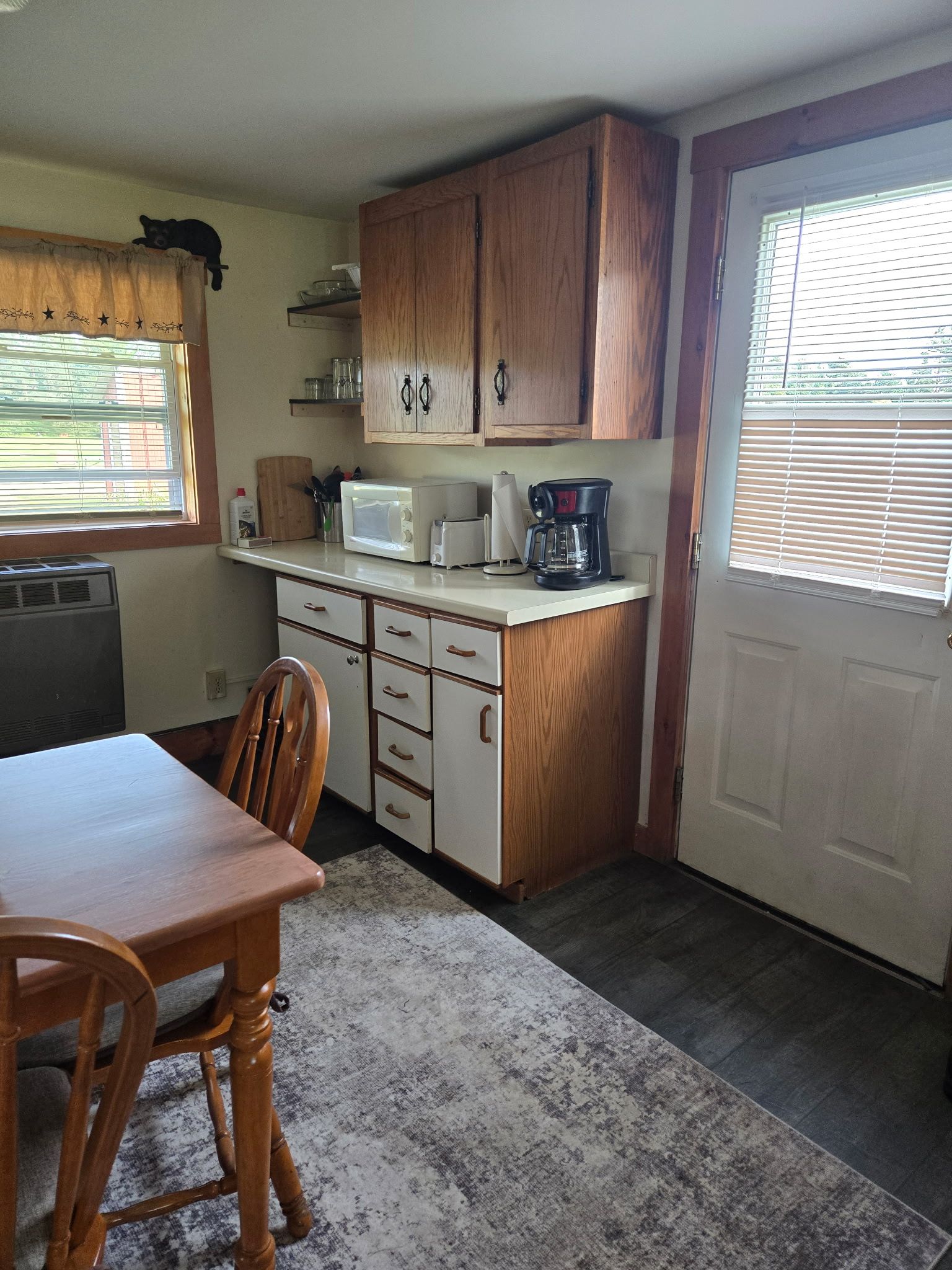 Small kitchen with wooden cabinets, a table, microwave, coffee maker, and a white door by a window.