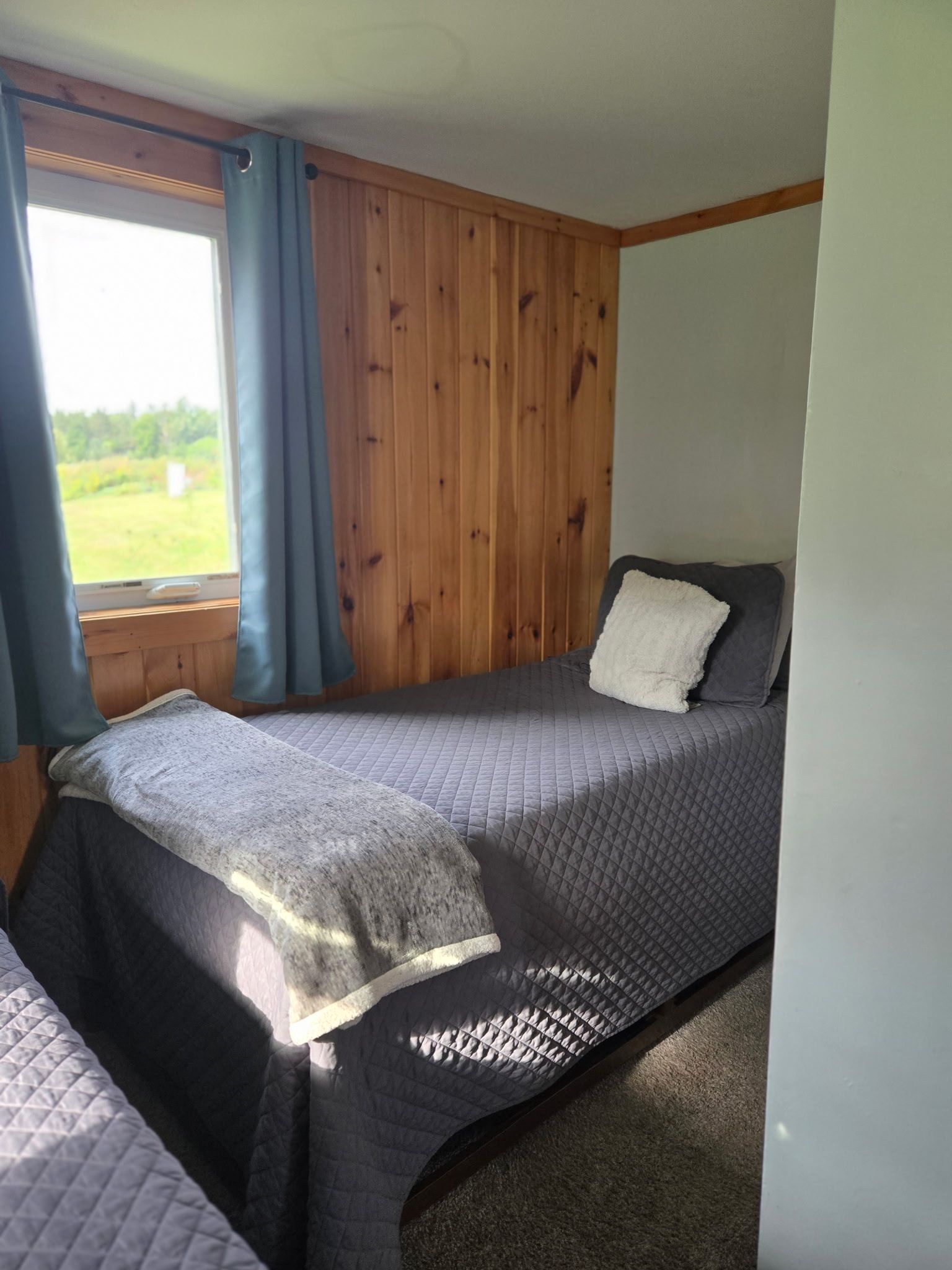 Small bedroom with a bed, dark gray bedding, wood-paneled wall, and a window with blue curtains.