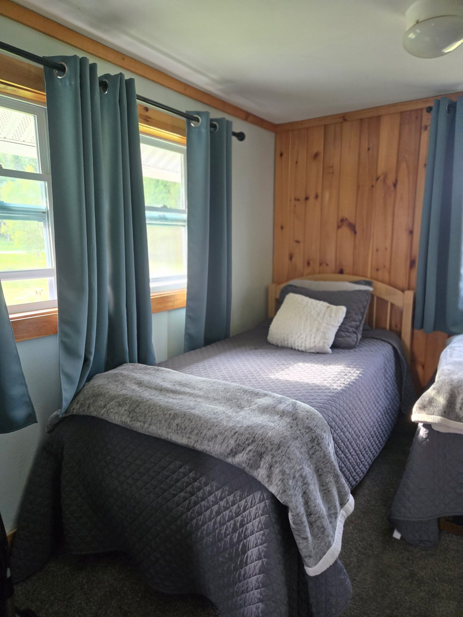 Small cozy bedroom with a single bed, gray bedding, blue curtains, and wood-paneled wall.