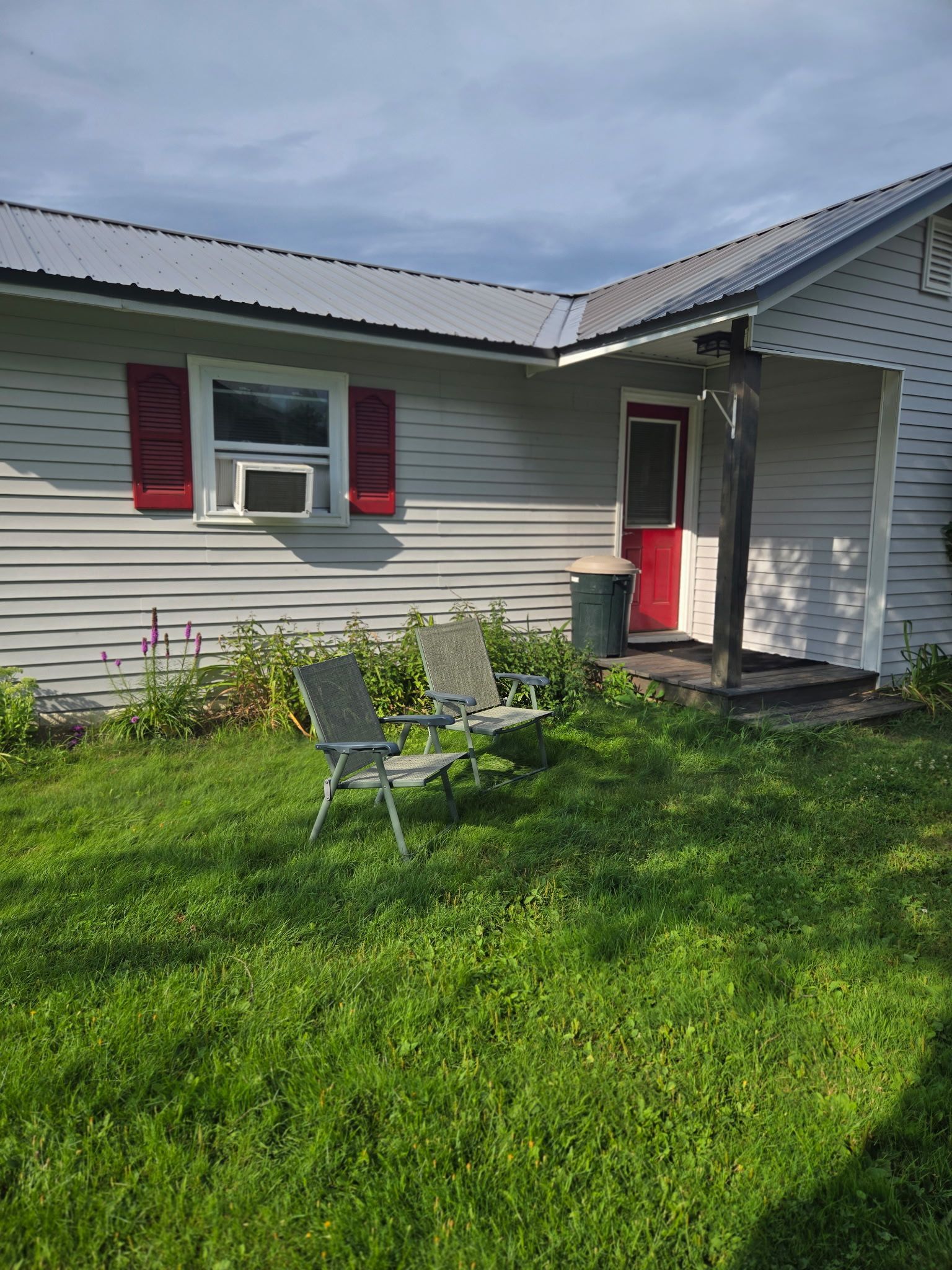 Single-story house with red shutters and door, small front porch, and lawn chairs on a green yard.