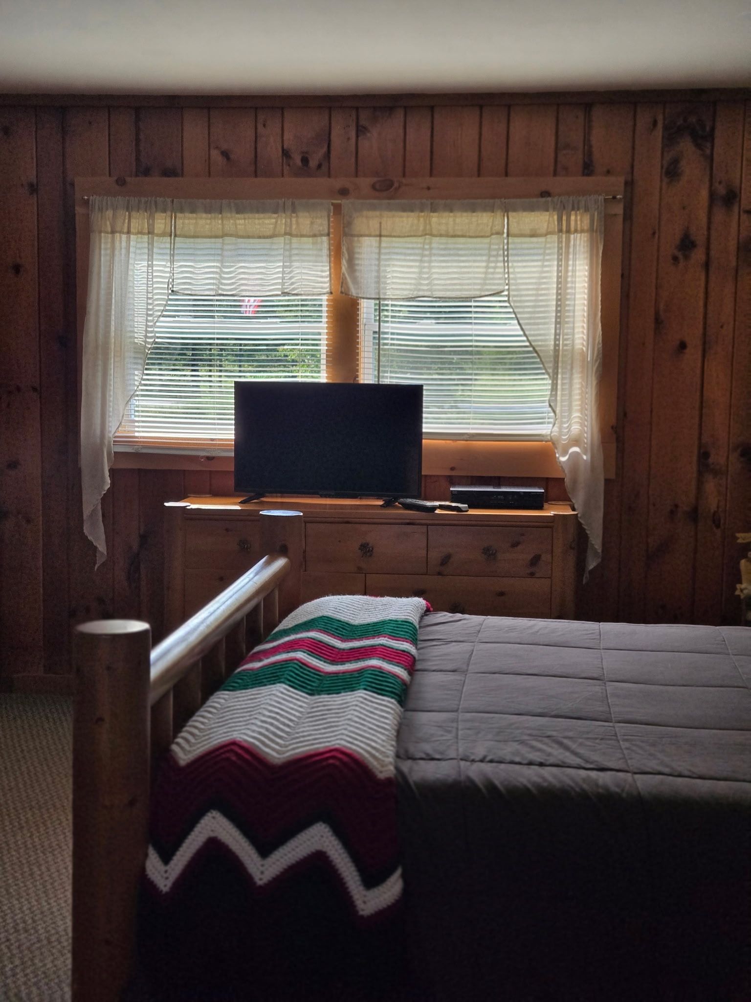Cozy wood-paneled bedroom with a bed, striped blanket, and a computer monitor by the window.