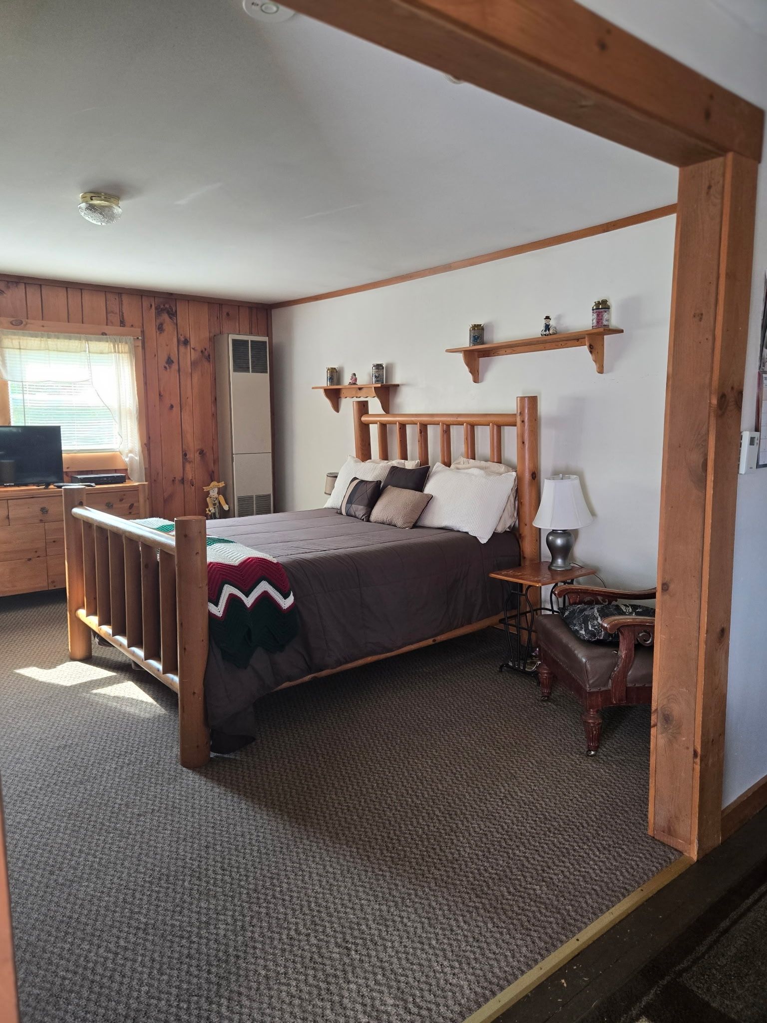 Rustic bedroom with wood trim, a large bed, patterned carpet, and a window with blinds.