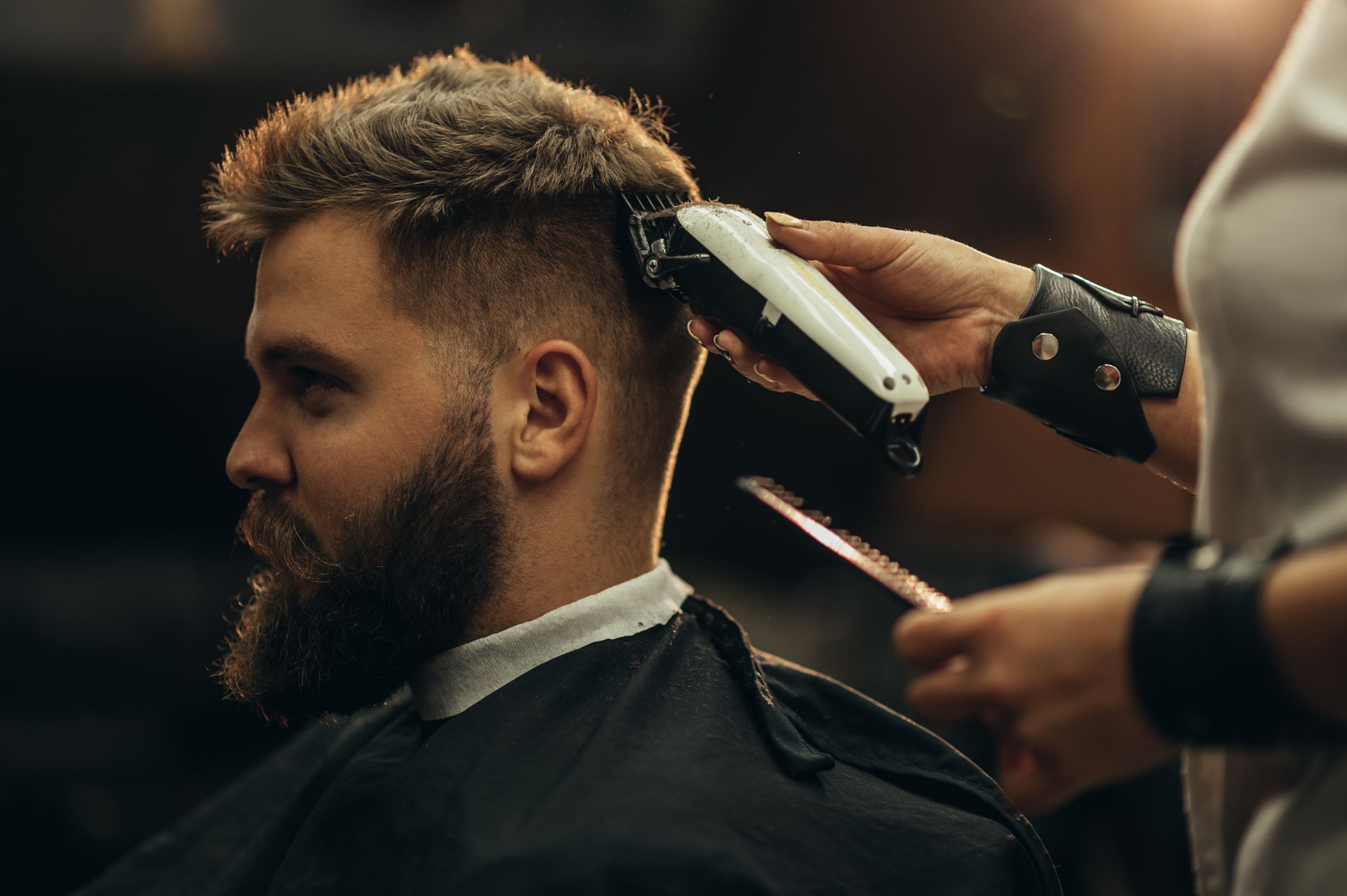 Barber trimming a bearded man's hair with clippers in a dimly lit salon