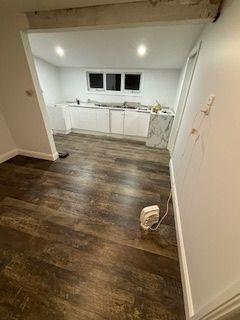 Newly renovated room with wood-look flooring and white walls, leading to a kitchen area with cabinets and a window.