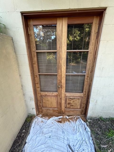 Wooden double doors with window panes and blinds, set into a white brick wall.