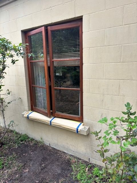 Wooden window, one panel open, on beige brick building with makeshift wooden sill.