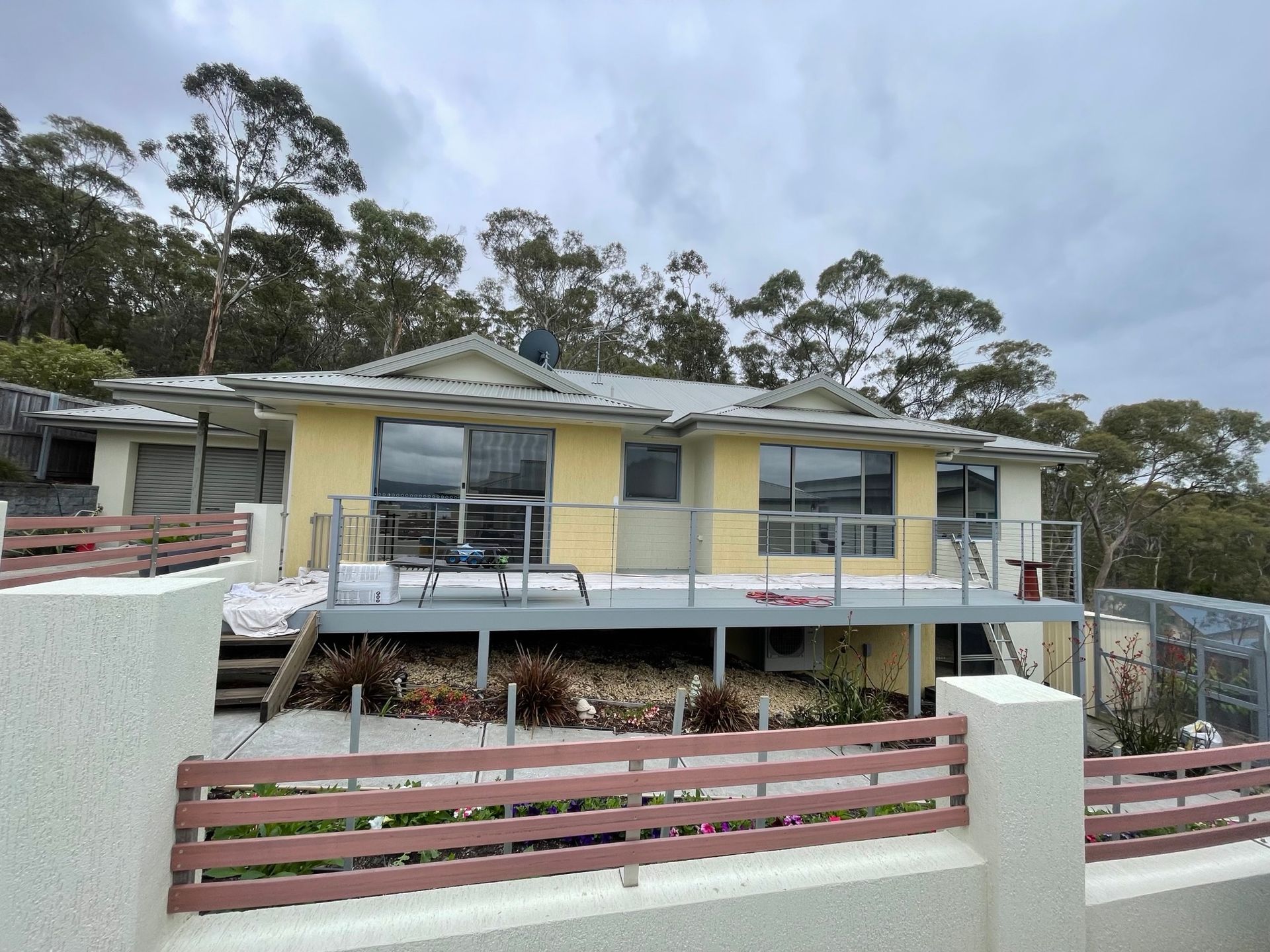 Yellow house with gray deck and roof, set on a hill, brown and white fence.