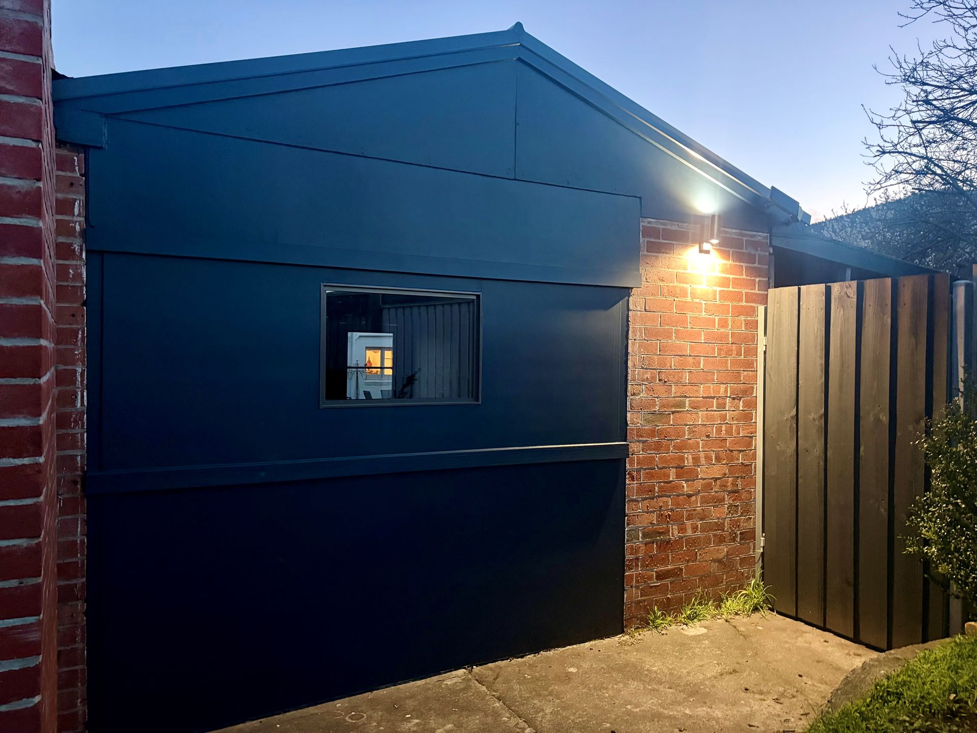 Dark blue exterior wall with a small window, brick corner, and lit porch light beside a wooden fence at dusk