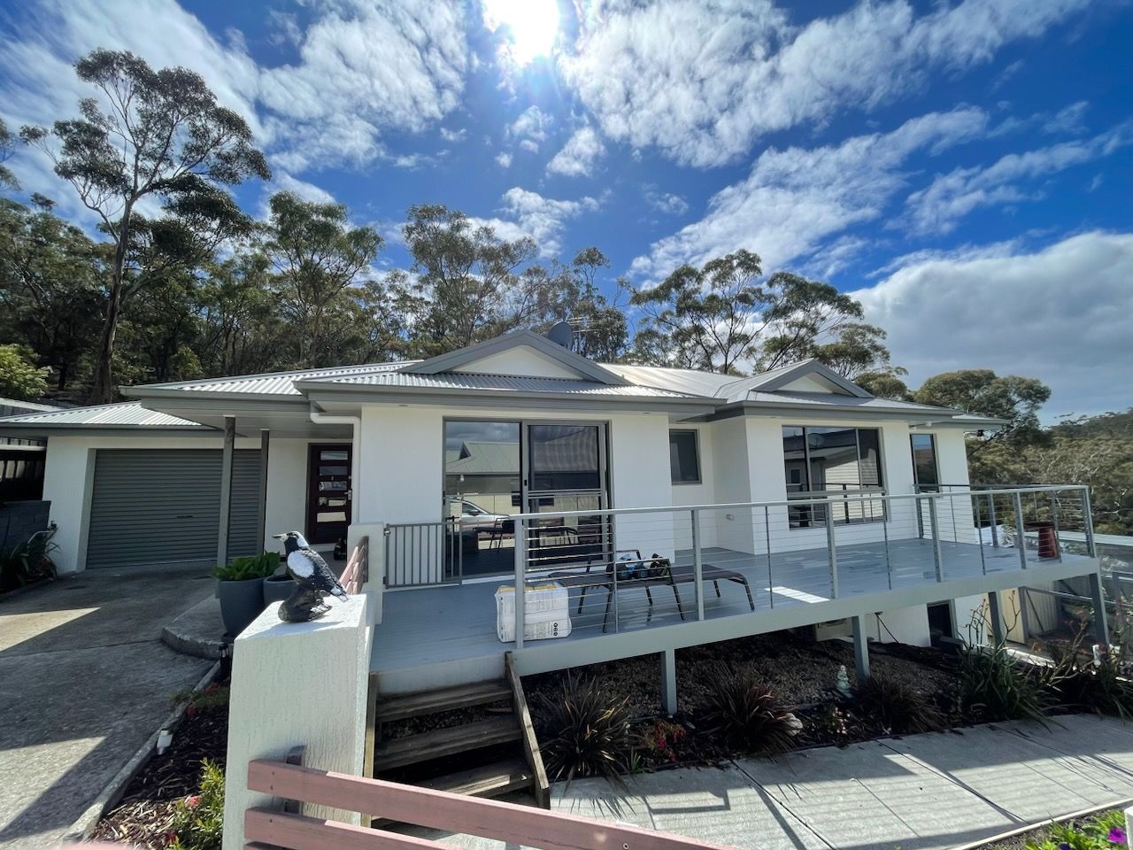 White house with a gray deck, overlooking a body of water, under a partly cloudy blue sky.