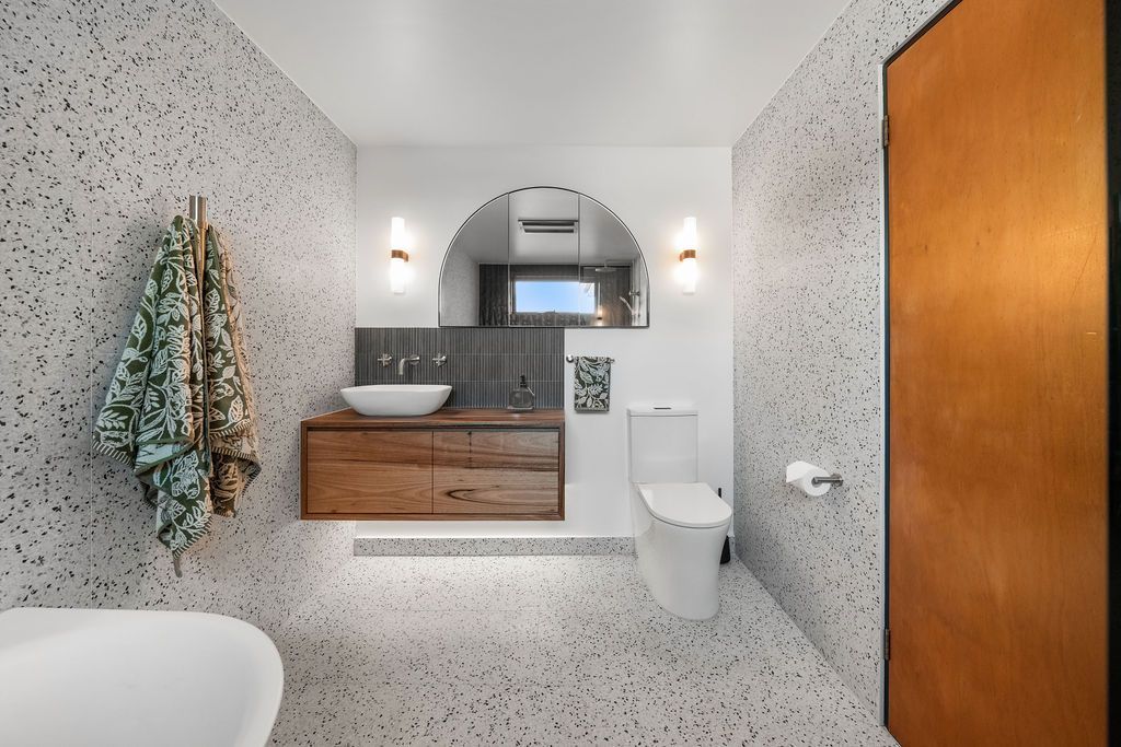 Modern bathroom with speckled tile, wood vanity, round mirror, and white toilet near a wooden door