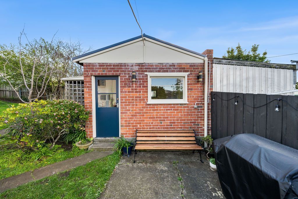 Small brick shed with blue door, front bench, gravel path, and black-covered object beside a fence.
