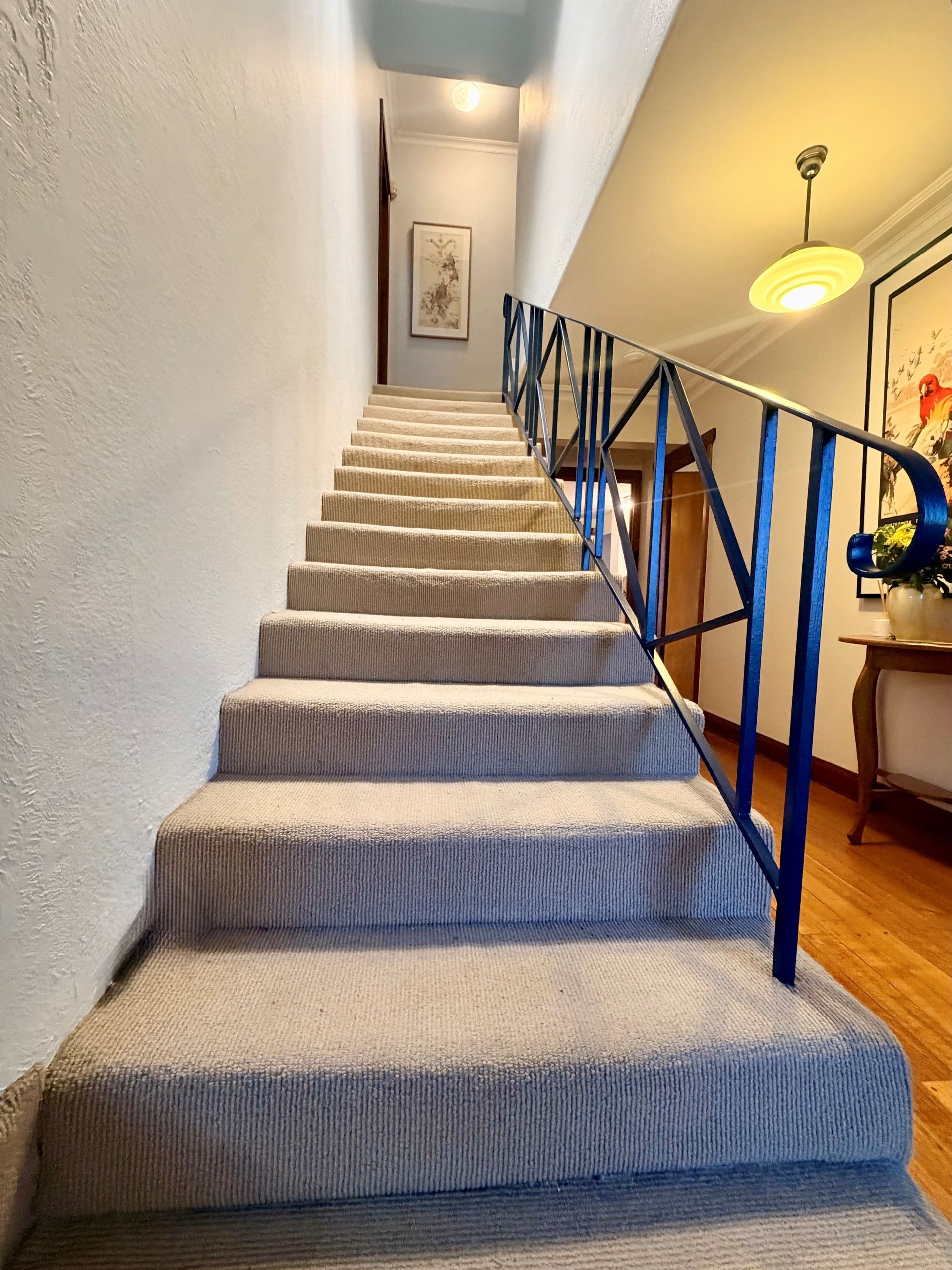 Carpeted staircase with blue railings in a narrow hallway, leading to an upstairs landing.
