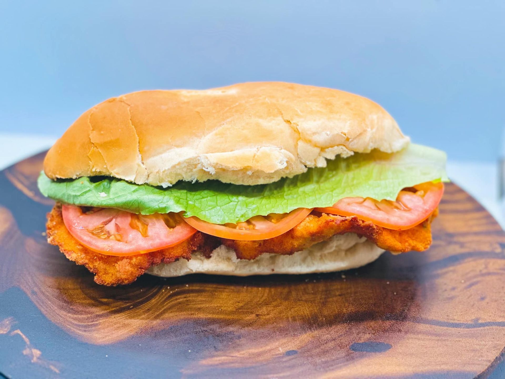 A chicken sandwich with lettuce , tomatoes and cheese on a bun on a wooden cutting board.