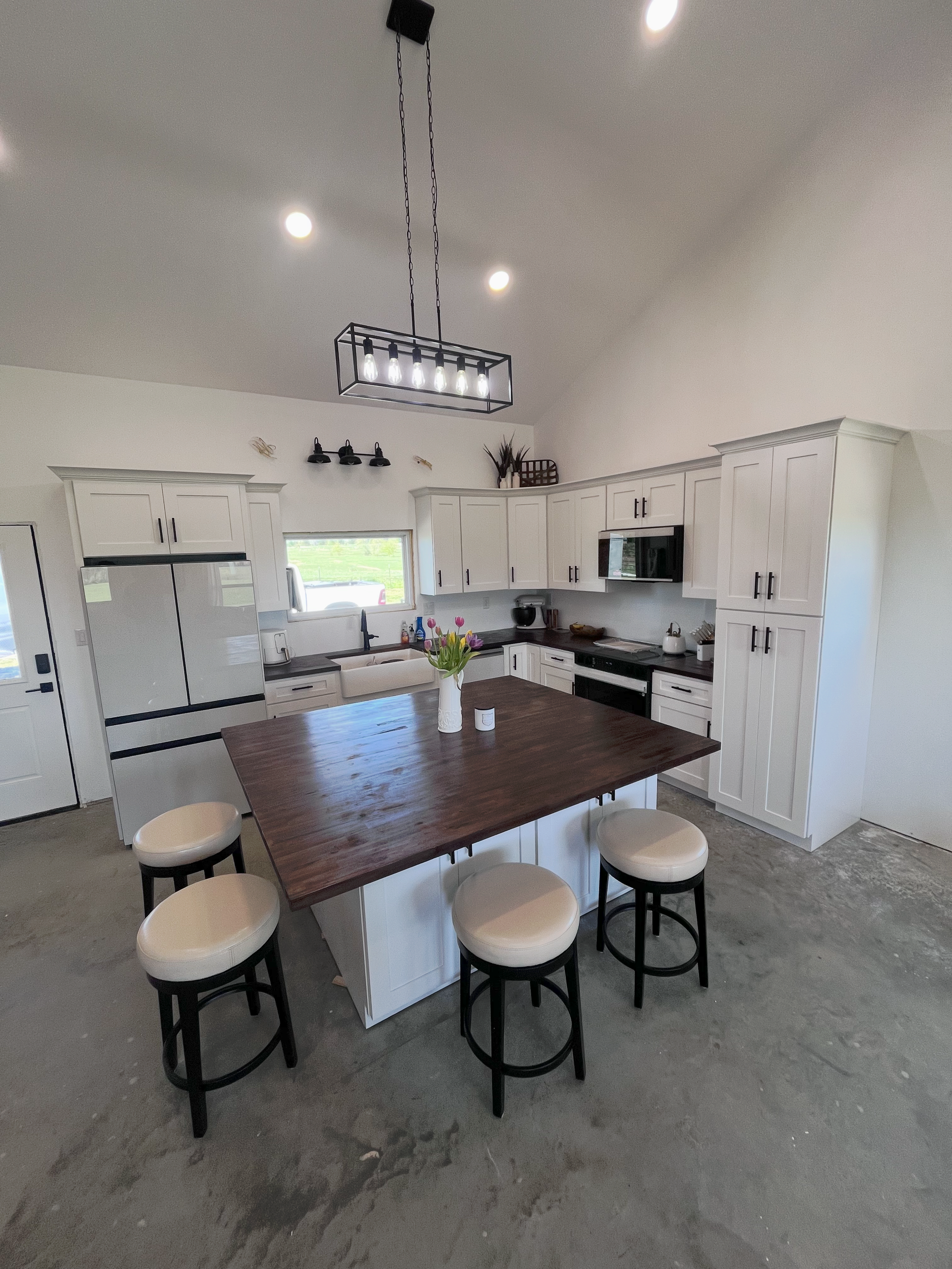 Bright modern kitchen with a large wood island, white cabinets, black stools, and a hanging light fixture