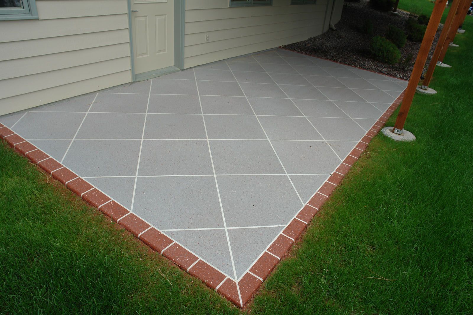 A grey concrete patio with a white diamond-grid pattern and a red brick border next to a house exterior.