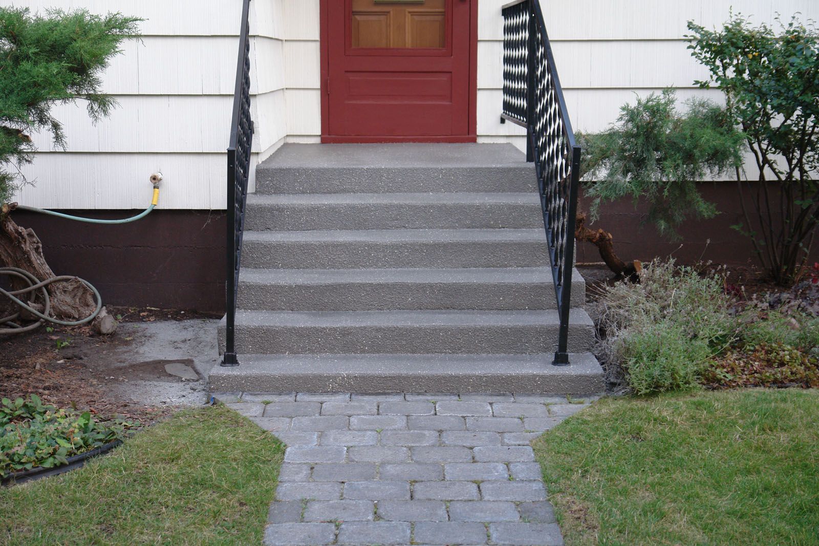 A red front door at the top of six concrete steps with black metal railings and a grey stone paver walkway.