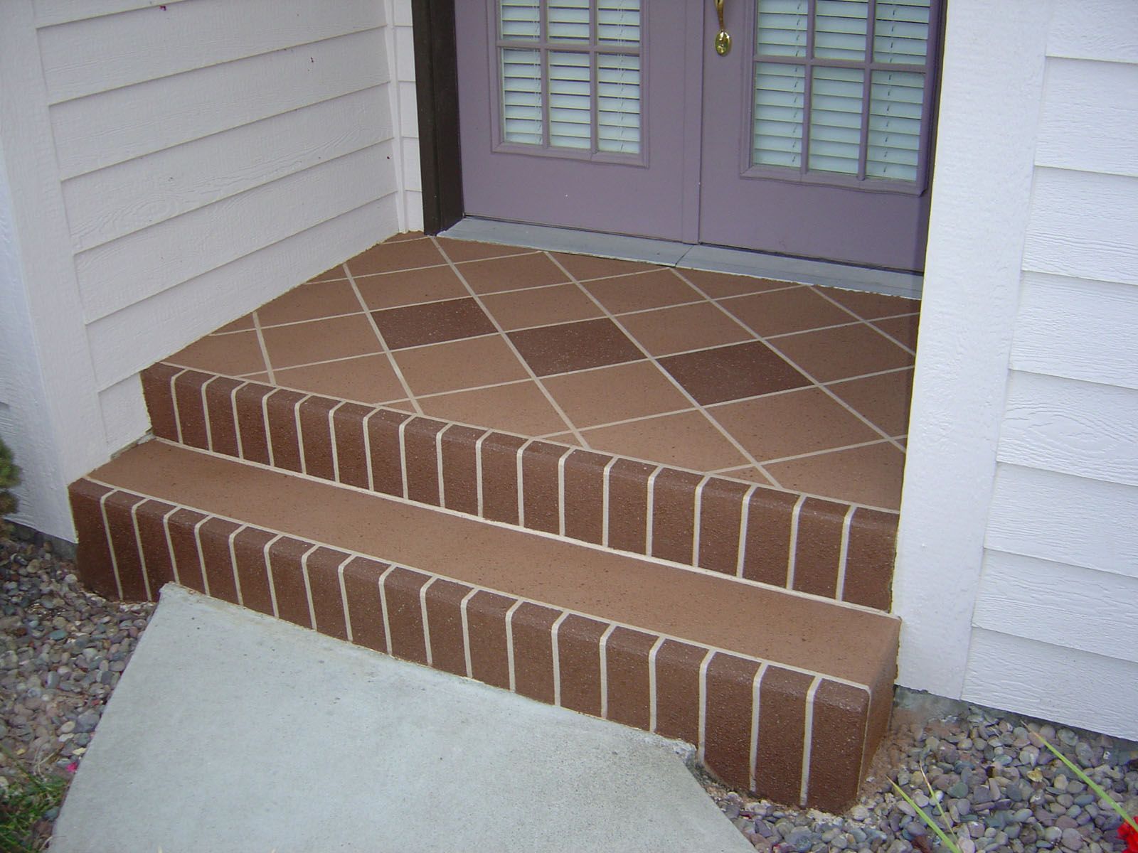 Two concrete steps leading to a house entrance, featuring a brown diamond-patterned top landing and brick-look risers.