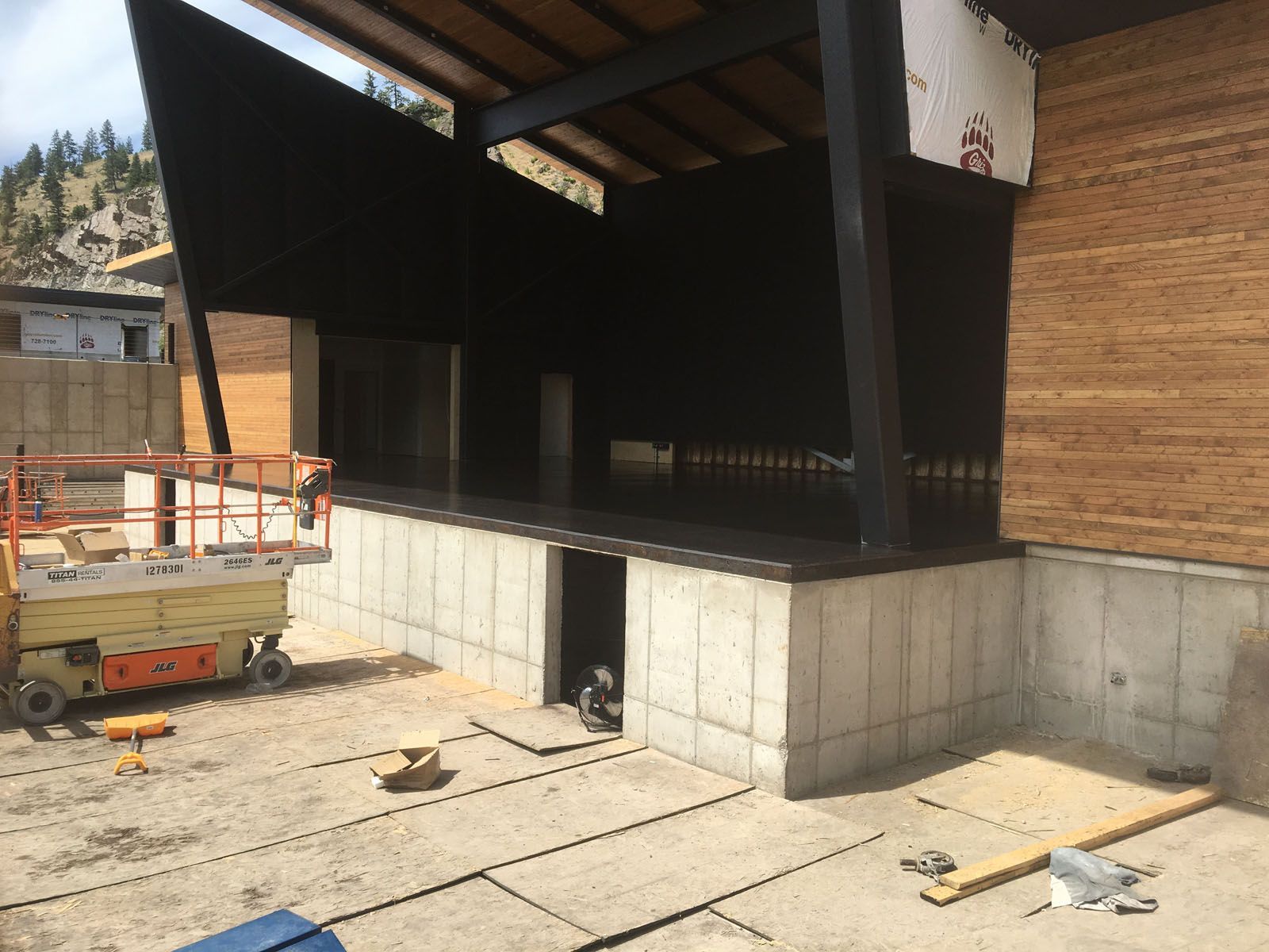 A modern outdoor stage structure under construction with wood-paneled walls, concrete foundations, and a scissor lift.