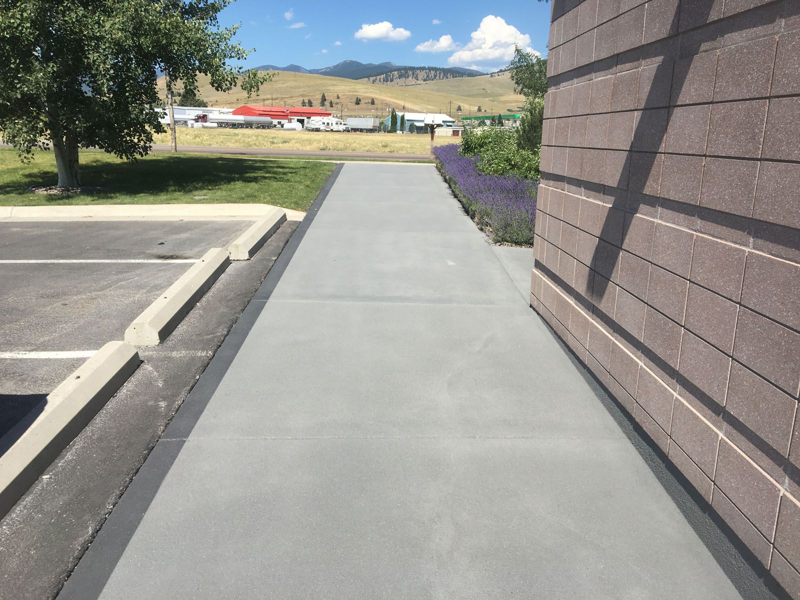 A paved sidewalk runs between a parking lot curb and a brick wall, leading toward trees and distant mountains.