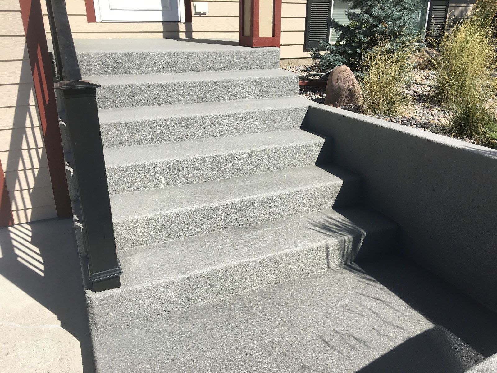 A set of outdoor concrete stairs with a speckled finish and a black metal handrail leading to a house entrance.