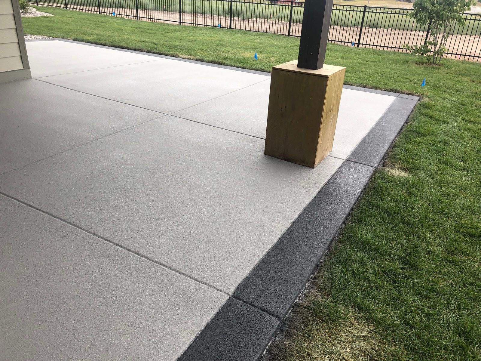 A concrete patio with a light gray center and dark gray border next to a lawn with a wooden support post.