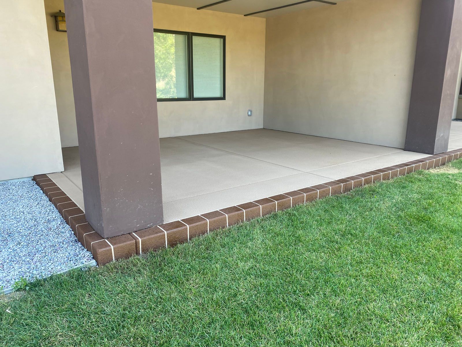 A covered concrete patio with a brown brick border, set against a house wall with a window, next to a grass lawn.
