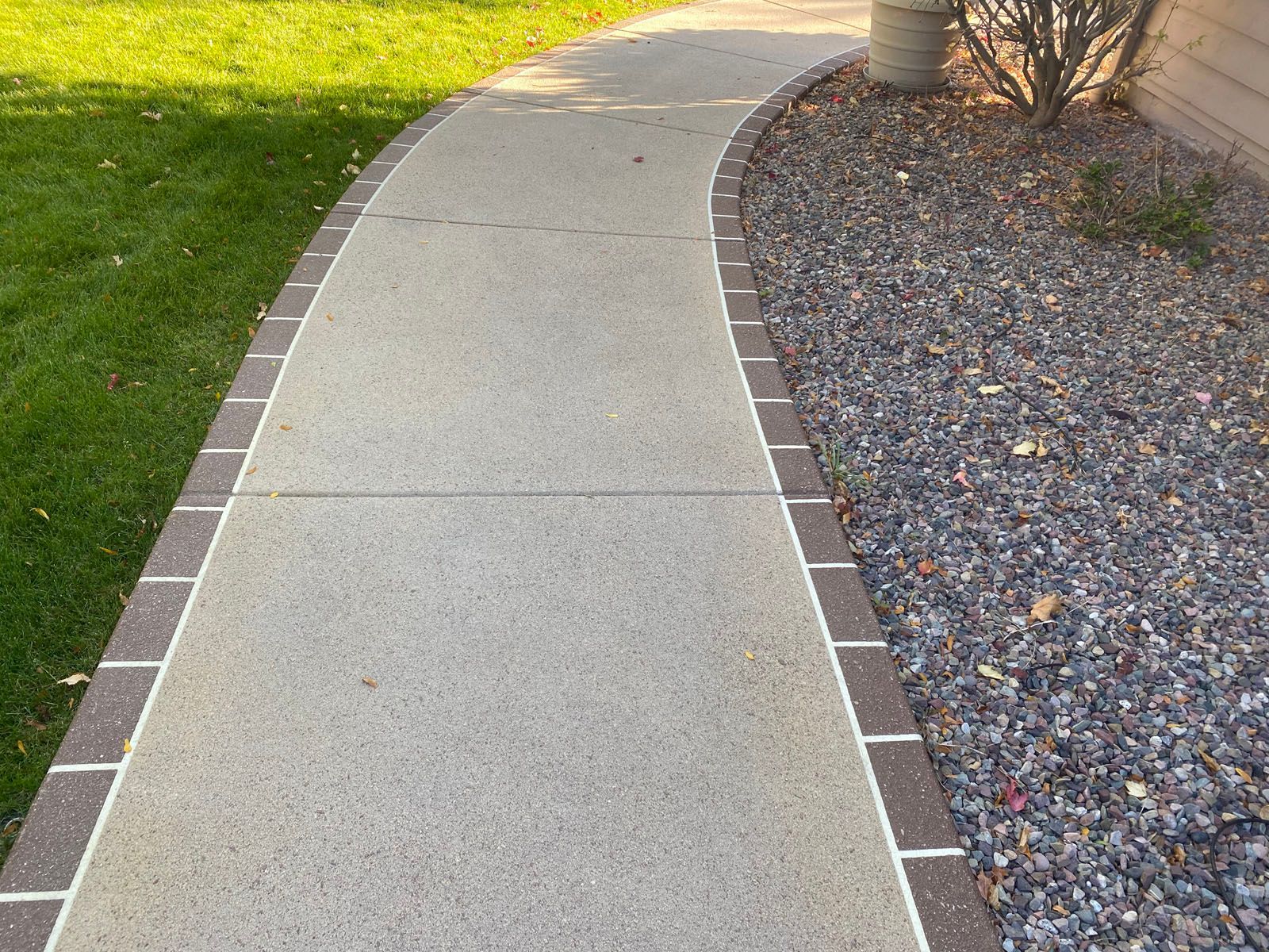 A curved concrete walkway bordered by bricks, with green grass on one side and a grey gravel bed on the other.
