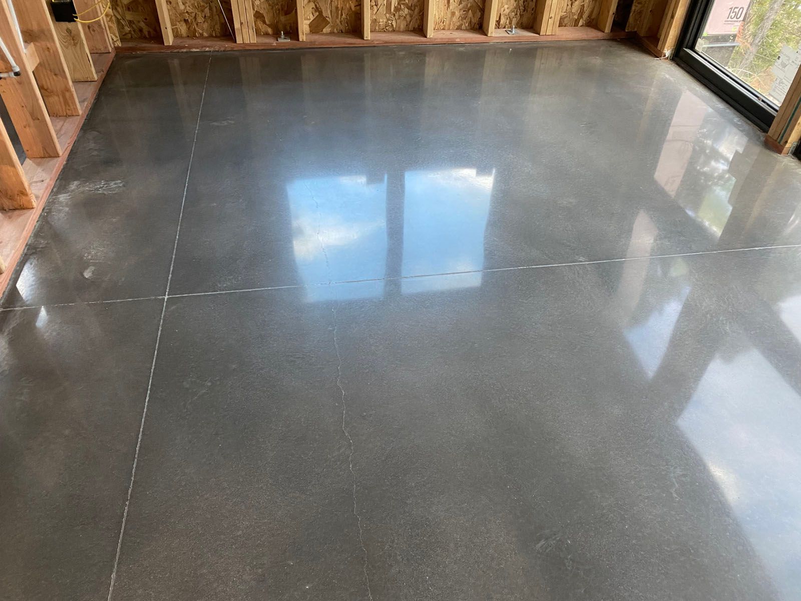 Polished gray concrete floor in a room under construction with wooden wall studs and a window reflecting the sky.