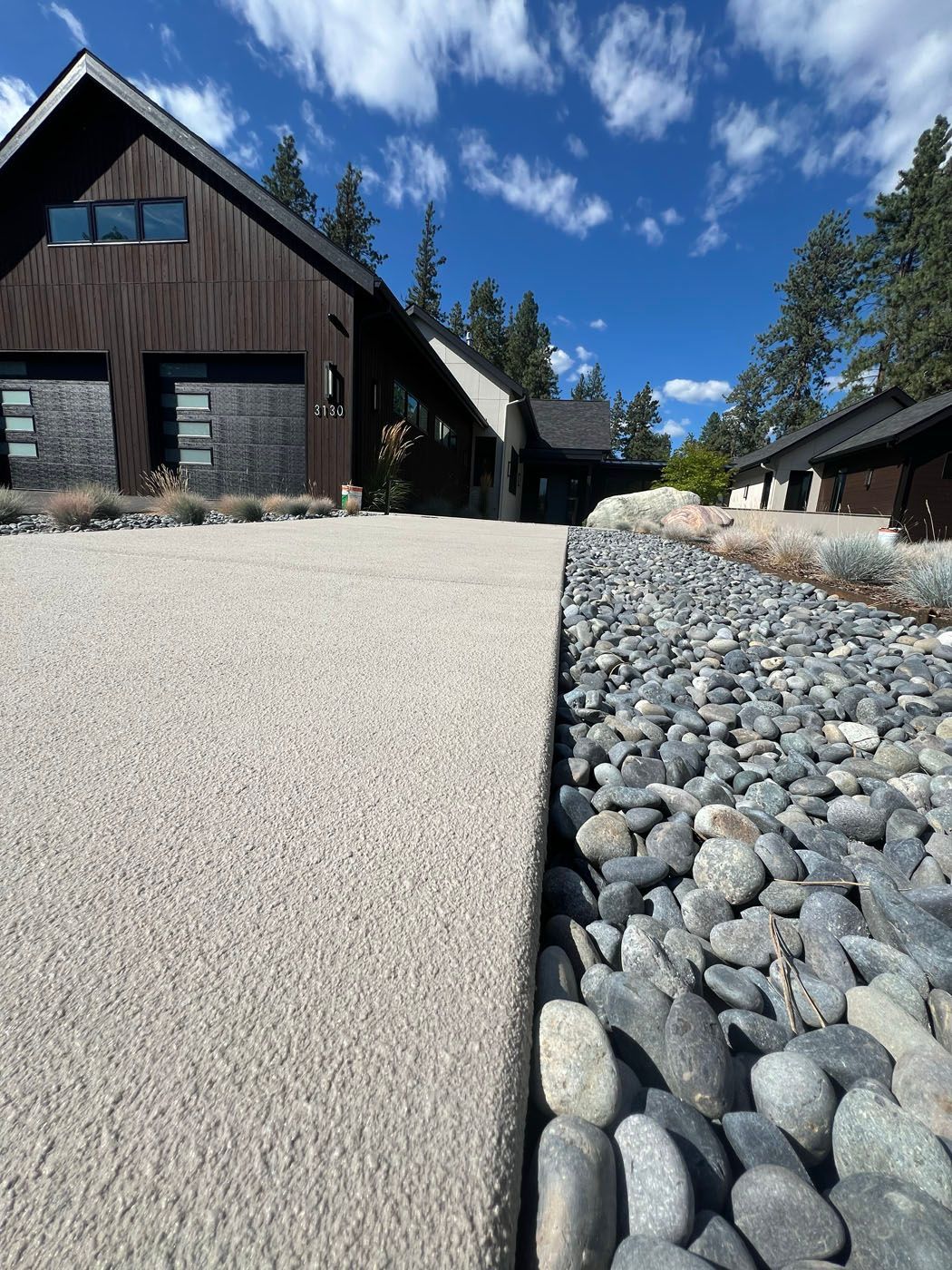 A concrete driveway edge runs alongside a decorative stone drainage strip leading to a dark modern house under a blue sky.