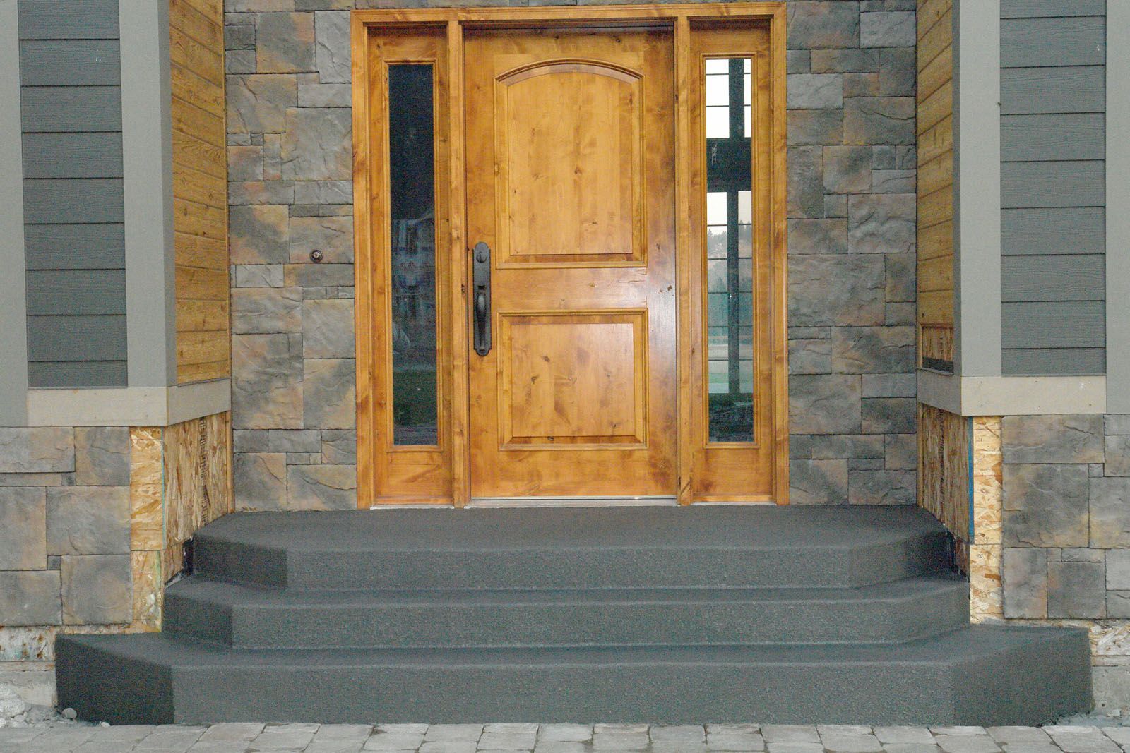 Wooden front door with glass side panels, set above three dark grey concrete steps, flanked by stone-patterned walls.