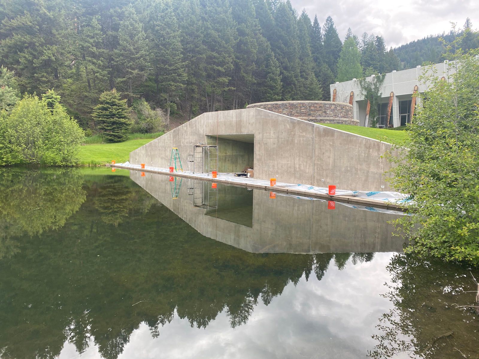 A concrete tunnel entrance sits at the edge of a pond, with a forested hillside and a building in the background.