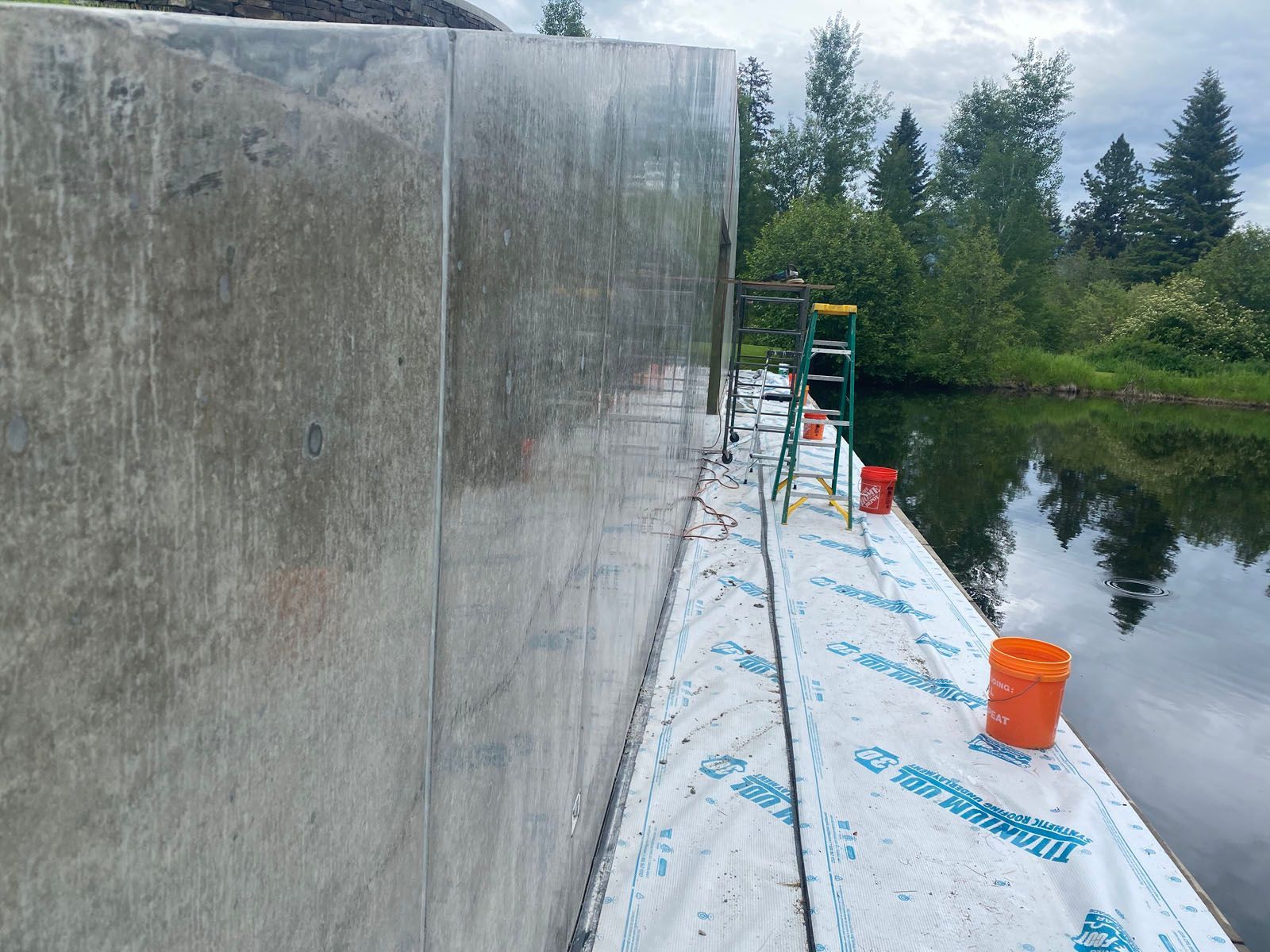 A tall concrete wall runs alongside a water-repellent barrier next to a body of water, with a ladder and orange buckets.