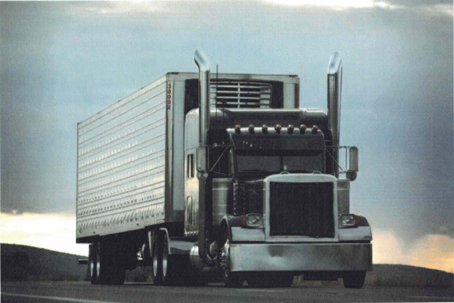 A large, silver semi-truck driving on a road under a cloudy sky.