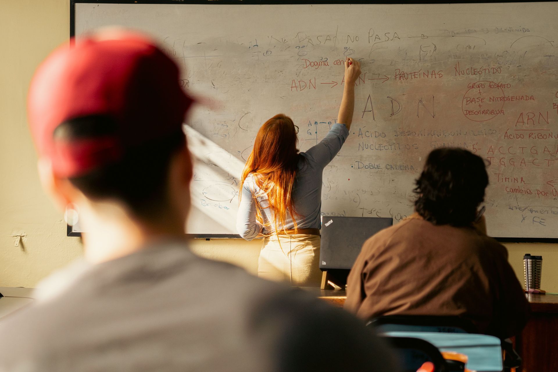 Students in a classroom, one writing on a whiteboard while others watch from desks