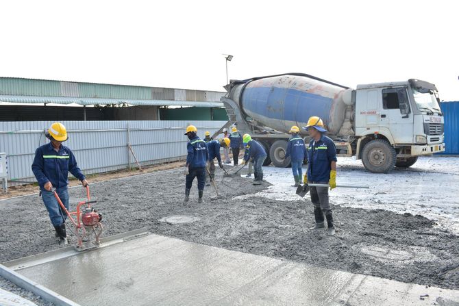 Construction workers paving a road beside a cement mixer truck and warehouse.