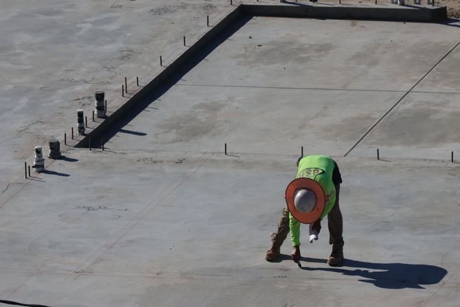Worker in a neon safety vest bending over on a concrete rooftop near a red coil or hose