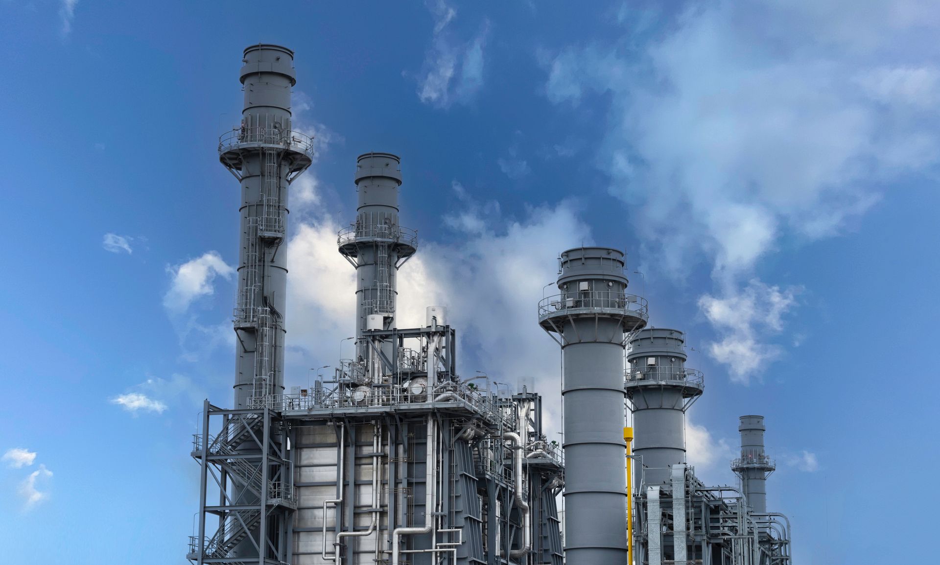 Natural Gas Plant with tall smokestacks and metal structures emitting steam against a blue sky with clouds.