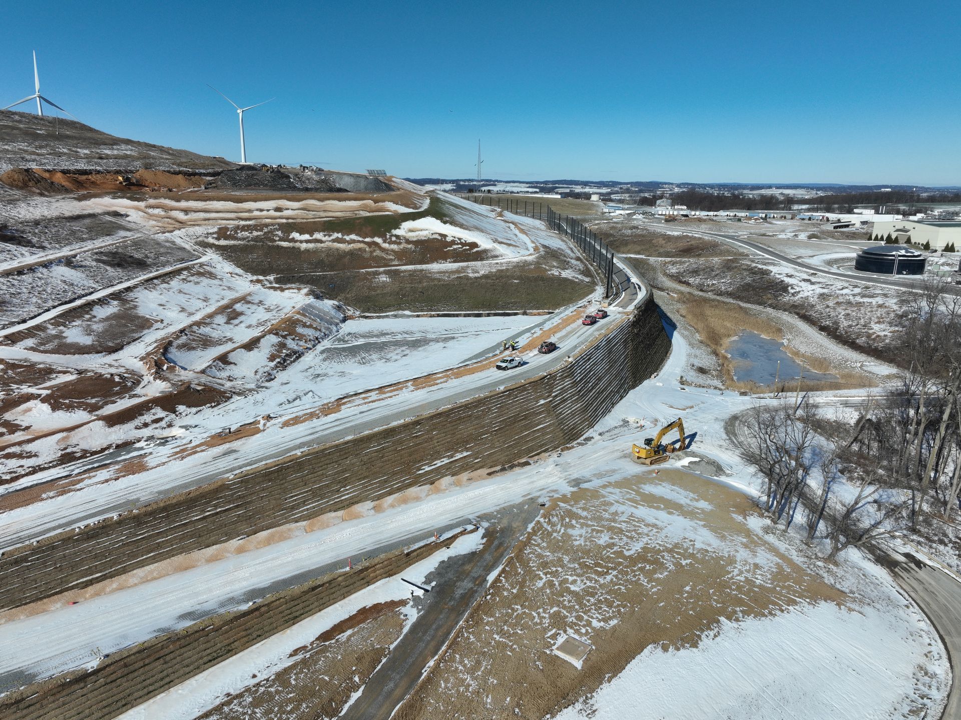 Winter aerial view of a landfill MSE berm under construction with active earthwork and wind turbines in the background.