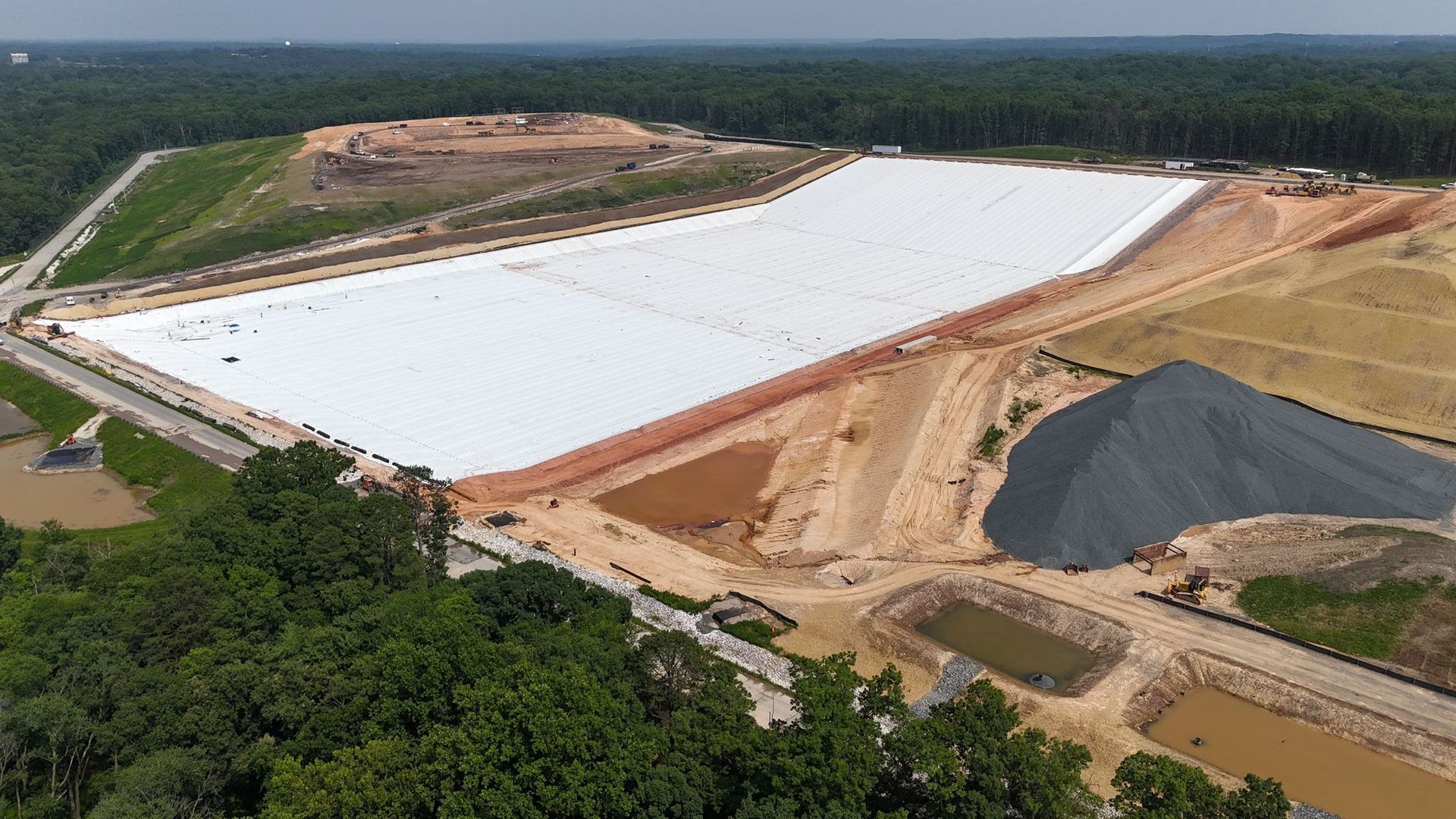 Aerial view of a newly lined landfill cell under construction, showing the white geomembrane liner, graded earthwork, access roads, and surrounding wooded areas